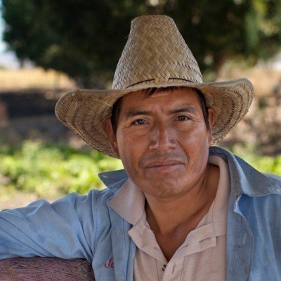 man in straw hat outside, wearing a light blue overshirt and white undershirt. the photo is close up on his face and he is outside