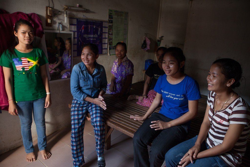 WIC community organizer Sot Sorphorng (center, right, blue shirt) with her sister Sot Pha (center, left, denim shirt) and four fellow garment workers sit together in the room they share. All six women sleep on the wooden platform pictured here. Photo by Evan Abramson.