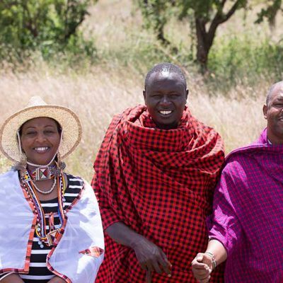 three women standing next to each other and smiling at the camera. the woman on the far left has a sunhat on. there are trees behind them.