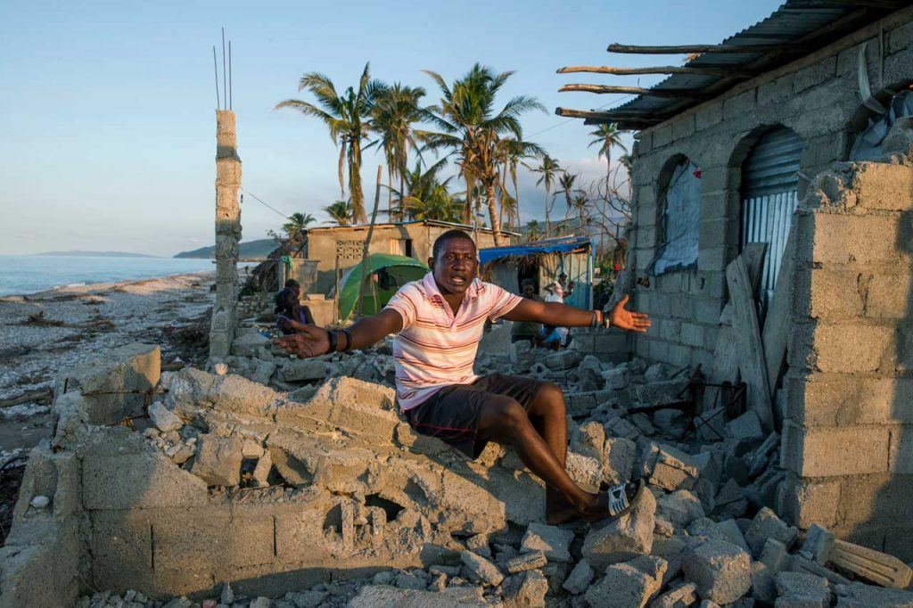 A man named Cidieu Jean Baptiste throws up his arms in frustration while sitting on top of the pile of rubble that used to be his home, which is very close to the ocean.