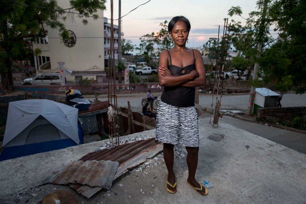 Françoise Inocent wearing a skirt and tank top with her arms crossed, standing at the site of what remains of her damaged home in Jérémie following Hurricane Matthew. Her home has a foundation but no roof or walls. There is a tent inside.