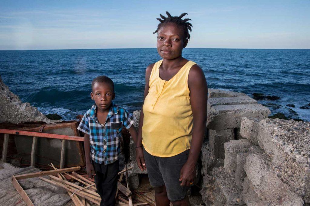 Gertie Mathurin standing with her young son Moise Henri wear serious expressions at the site of their damaged home in Jérémie. The walls and roof of the home are gone, and it is right on the coast. Gertie has received support from AJWS grantee SOFA. Photograph by Jonathan Torgovnik