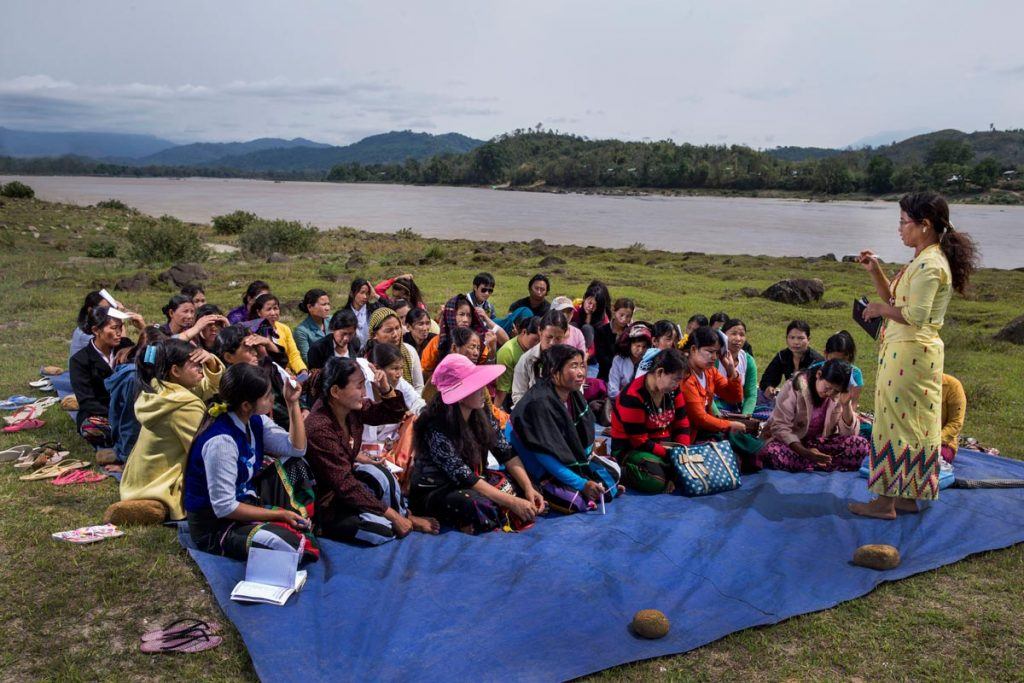 Women sit on a tarp near the river and participate in a community meeting with MRJ. 