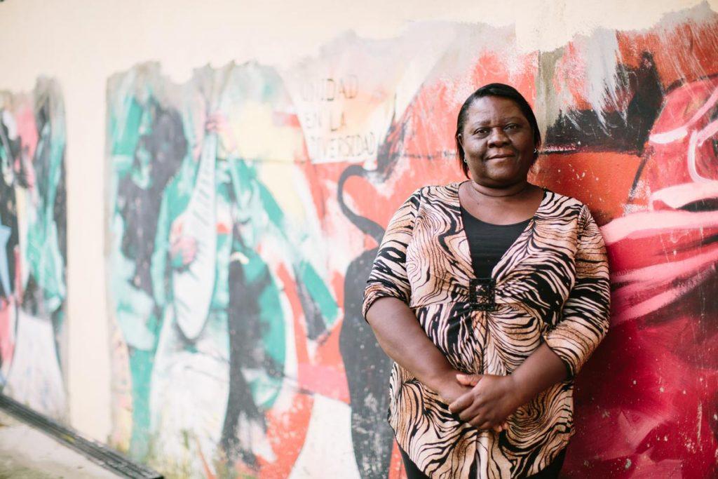 Germania Sevito, (standing in front of a colorful mural with her hands together, smiling softly) a Dominican of Haitian descent from Yacó Batey west of Santo Domingo, fought for access to her birth certificate for seven years. AJWS grantee Reconoci.do has helped her rise above the persecution she has faced because of her Haitian heritage. Photograph by Christine Han 