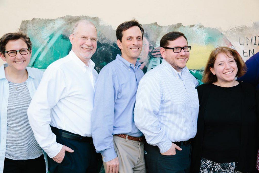 Rabbi Bruce Elder (center, wearing a blue shirt and smiling) standing in front of a colorful mural with colleagues Rabbis Sharon Kleinbaum (wearing glasses and a striped shirt), Sam Gordon (wearing a white shrit and glasses), Daniel Stein (in a light blue shirt and glasses) and Lauren Herrmann (wearing a black shirt) outside the offices of Reconoci.do, where the fellows learned about the Dominican citizenship crisis from people who have been affected by it. 