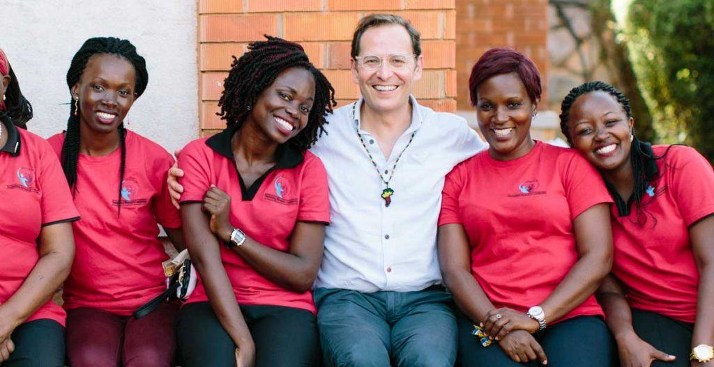 Robert Bank in the center sitting and smiling with four other women from the Ugandan organization MEMPROW. 