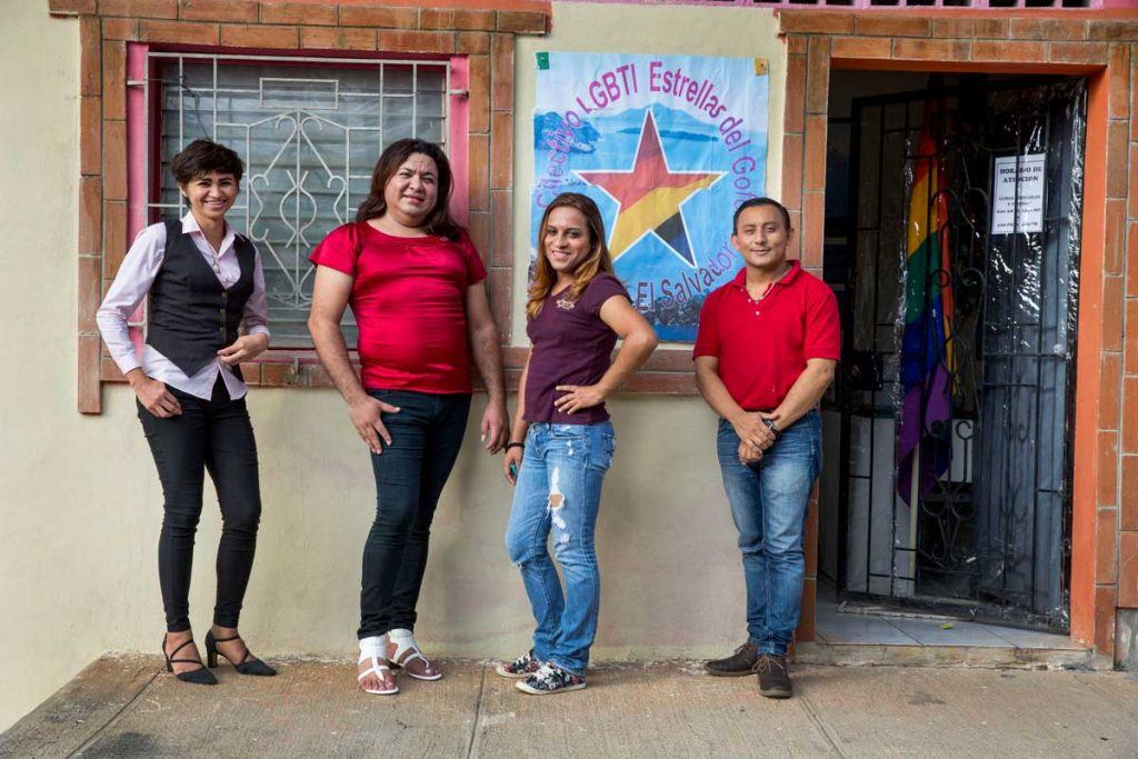 Four leaders of Estrellas del Golfo smile and pose outside their office in La Union, El Salvador. There is a large sign with a rainbow star and the name of their organization on the wall. Photo by Jonathan Torgovnik