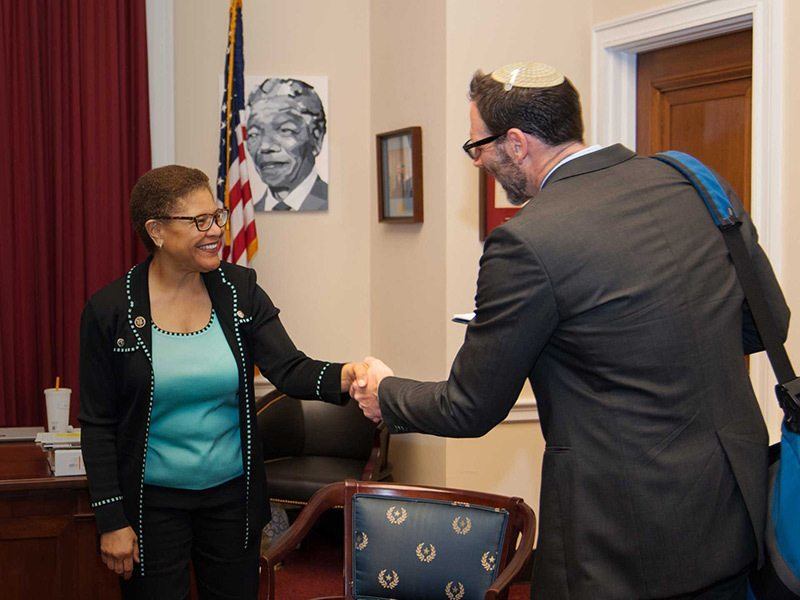 three people smiling in a government office. a woman and man are looking at each other and shaking hands. the man is wearing a suit and yarmulke. the woman is wearing a blazer and blue blouse.