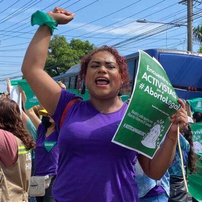 woman at a protest, wearing a purple tshirt, holding a green sign with a solidarity fist in the air