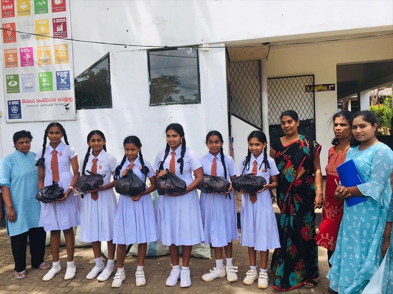 Group of students in white dresses standing alongside a few adults in front of their school.