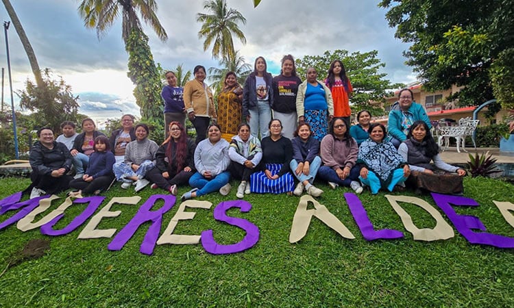 group of about twenty people standing outside under some palm trees, with "Mujeres ALDEA" spelled out in front of them on the grass in large purple and yellow letters