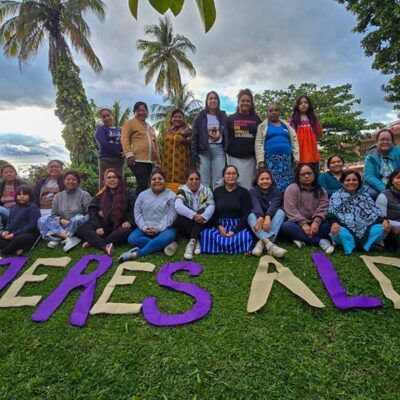 group of about twenty people standing outside under some palm trees, with "Mujeres ALDEA" spelled out in front of them on the grass in large purple and yellow letters