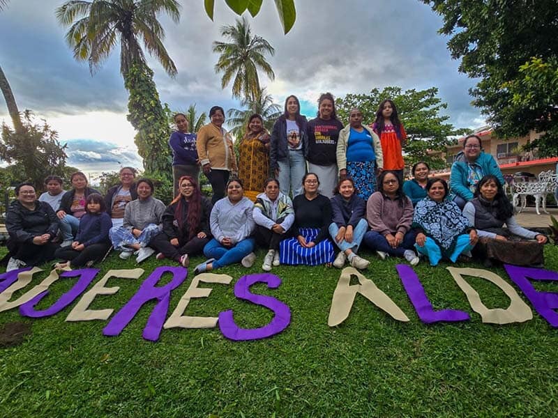 group of about twenty people standing outside under some palm trees, with "Mujeres ALDEA" spelled out in front of them on the grass in large purple and yellow letters