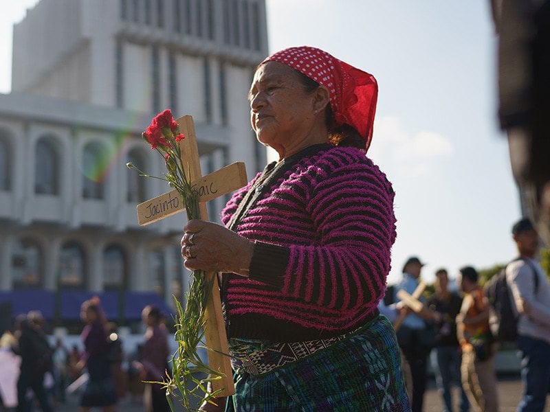 woman wearing a red striped shirt and red bandana standing up at the march, holding a wooden cross and red rose