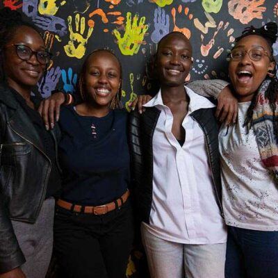 group of young people smiling at the camera in front of a wall painted with colorful handprints