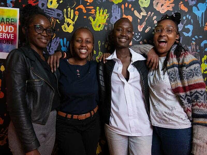 group of young people smiling at the camera in front of a wall painted with colorful handprints