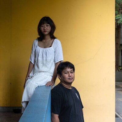 two people with yellow wall behind them, looking at camera