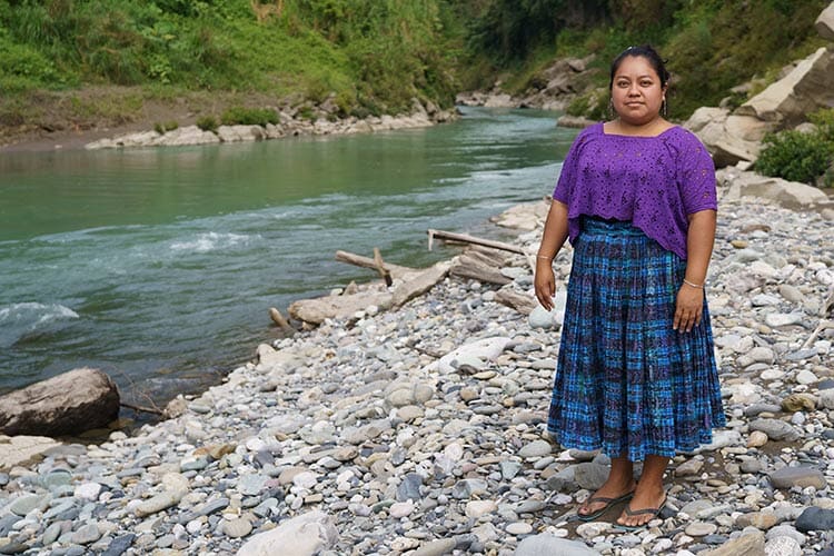 woman standing by a river wearing a blue skirt and purple top, looking at the camera