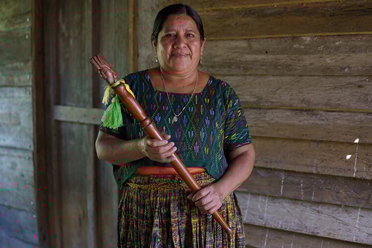 woman holding staff, wearing dress, looking at camera. in front of wooden wall