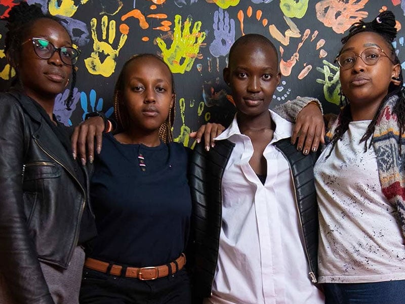 four young people standing together, arms around each other and looking at the camera