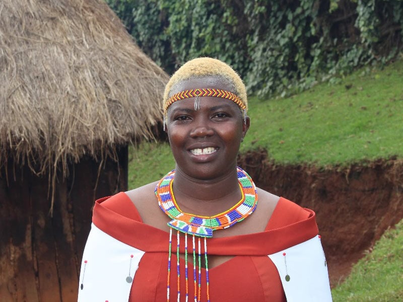 woman wearing red top and beaded accessories behind grass and a straw roof