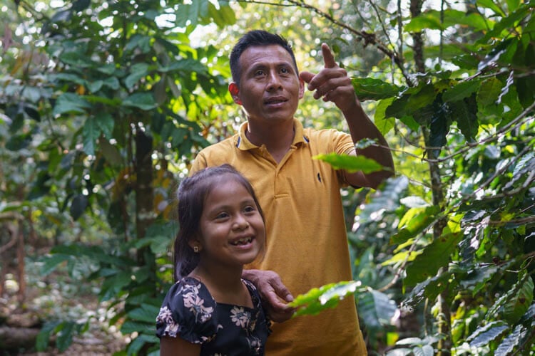 man in green collared shirt with young girl smiling, among lush greenery outside