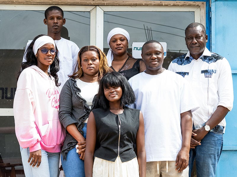 seven people standing outside in front of a building with a window and blue wall