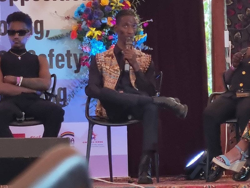 three people seated in chairs on a stage, the one in the middle speaking into a mic